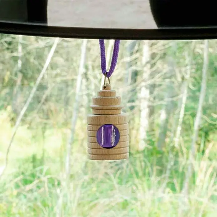 Ambientador de coche para colgar - Lavanda, 8 sem. - Un ambientador de coche de madera con un detalle púrpura cuelga del espejo retrovisor, proporcionando una fragancia agradable al conducir.