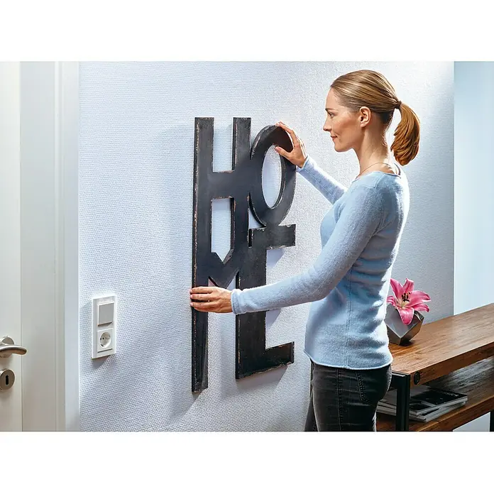 Tesa Clavo adhesivo - Blanco, Carga soportada: 2 kg, 2 ud., Paredes pintadas - Mujer cuelga un gran elemento decorativo negro con la palabra "HOME" en una pared blanca en un interior.