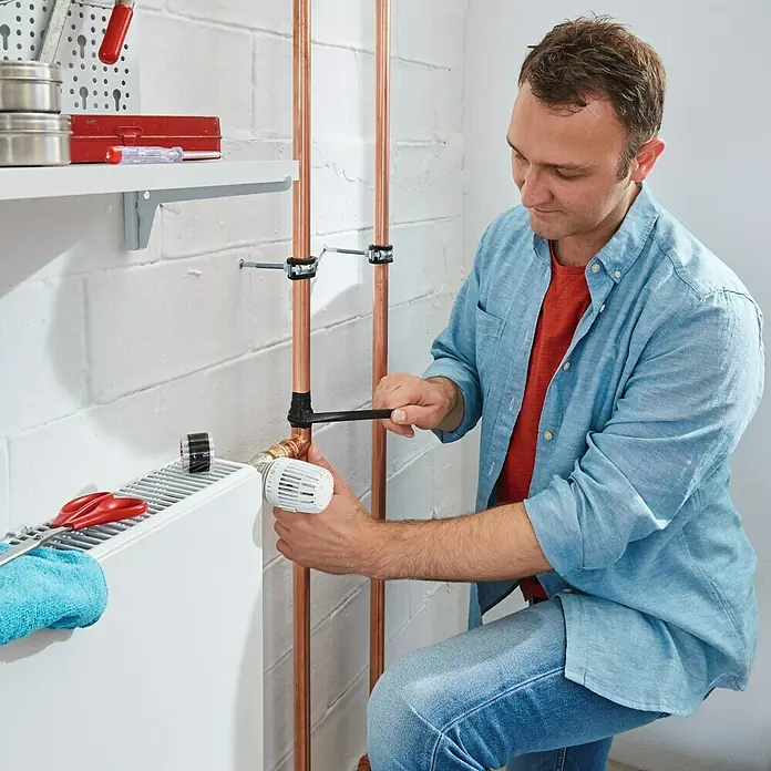 Tesa Cinta de reparación Extreme Repair - Transparente, 2,5 m x 19 mm - Un hombre está reparando las tuberías de cobre de un radiador con una llave inglesa. Se ven herramientas y un estante en la pared.
