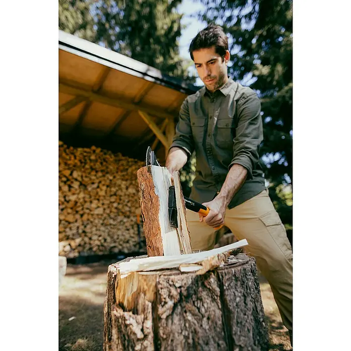 Fiskars X-series Universalaxt X14 - Länge: 35,5 cm - Ein Mann spaltet Holz mit einer Fiskars-Axt auf einem Baumstumpf. Die Axt ist schwarz und orange. Im Hintergrund befindet sich ein Holzschuppen.