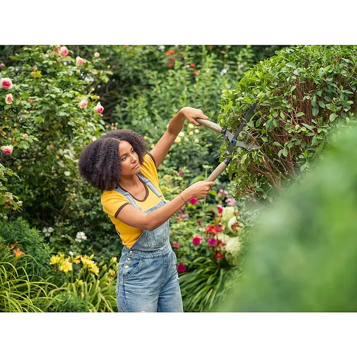 Gardena Tijeras de podar manuales NatureCut - Largo: 580 mm - Una joven con cabello afro, en overol, poda un seto con tijeras de jardín en un jardín floreciente.