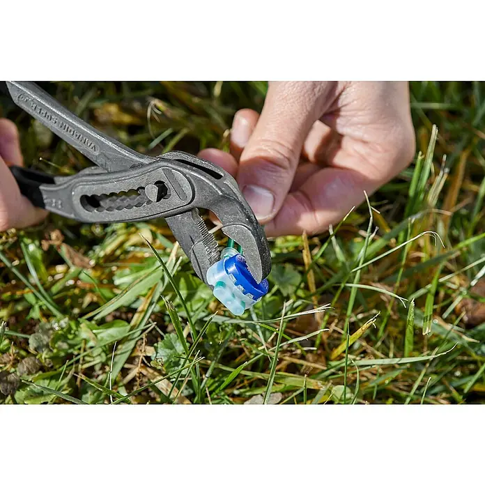 Gardena Conector clip para robot cortacésped R40LI - Específico para: Robot cortacésped Gardena - Un terminal de cable eléctrico multiconector se engarza con unos alicates contra un fondo de hierba, asegurando una conexión segura.