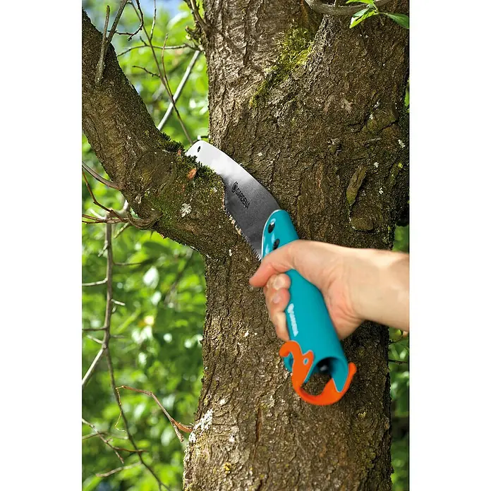 Gardena Combisystem Sierra de podar manual 300PP - Largo: 556 mm, Plástico, Plateado, Turquesa - Primer plano de una mano usando una sierra de mano Gardena para cortar una rama de un árbol. La sierra tiene un mango de color verde azulado.