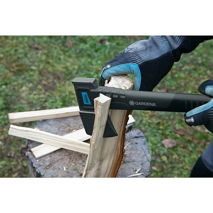 Gardena Universalbeil 900 B - Länge: 34 cm - Eine Gardena Spaltaxt wird zum Spalten von Holz auf einem Baumstumpf verwendet, mit Holzscheiten im Hintergrund. Die Axt ist schwarz mit blauen Akzenten.