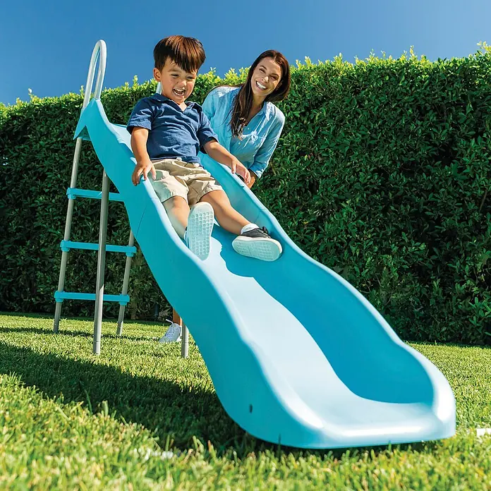 Intex Rutsche - L x B x H: 251 x 84 x 147 cm, Kunststoff, Himmelblau/Grau - Ein Kind saust auf einer blauen Kinderrutsche im Garten hinunter, während eine Frau lächelt. Die Rutsche hat eine Leiter für den Aufstieg.