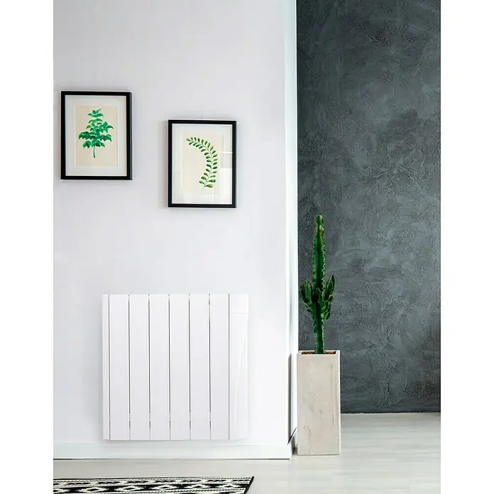 Voltomat HEATING Emisor térmico VOLT - 750 W, Blanco, 9,8 x 58,5 x 57,5 cm, Fluido - Radiador blanco debajo de dos impresiones artísticas botánicas, adyacente a un cactus alto contra una pared gris.