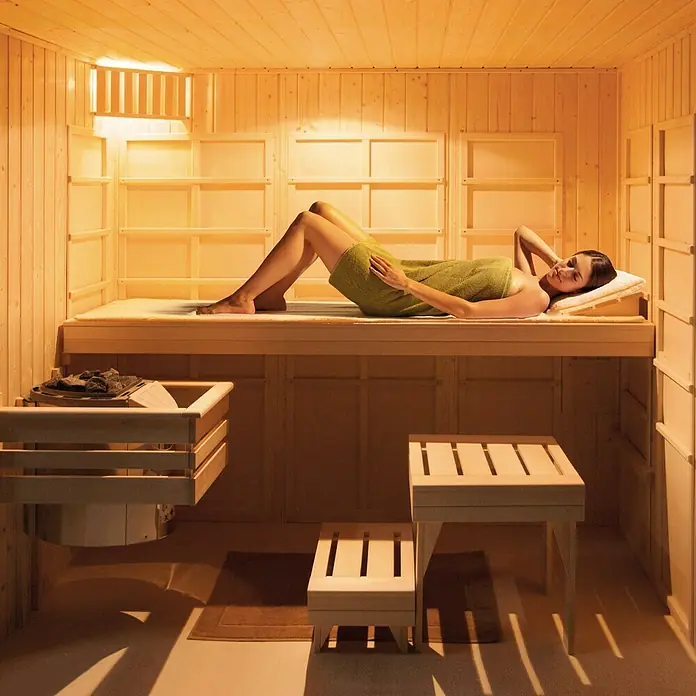 Weka Infrarot-Sauna-Kombikabine Uppsala - Therm-Flächenstrahler + 3,6 kW Bio-Saunaofen, 121 x 212 x 199 cm - Frau entspannt auf einer Holzliege in einer Sauna, die mit hellen Holzpaneelen verkleidet ist und ein warmes, einladendes Ambiente bietet.