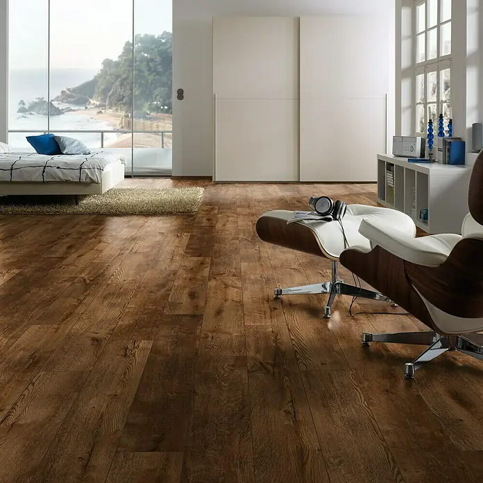LOGOCLIC Ambienta Laminatmuster Eiche Mirandola - 296 x 195 x 1 mm, Landhausdiele - Das Bild zeigt einen Raum mit einem dunklen Holzboden, einem Sessel, einem Bett und einem großen Fenster mit Meerblick.