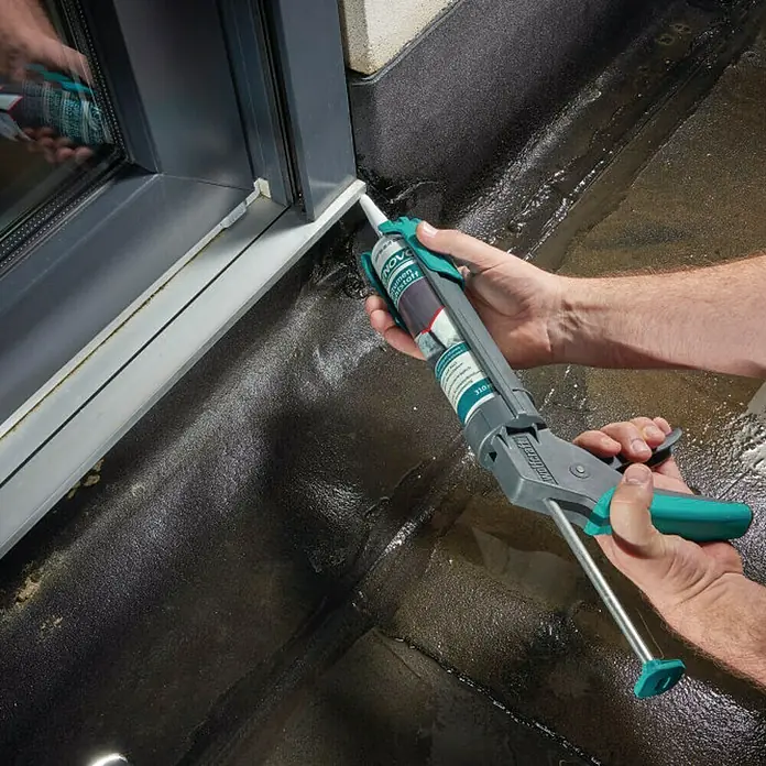 Wolfcraft Pistola de silicona MG 200 - Apto para: Silicona y cartuchos - Artesano utiliza una pistola de calafateo para sellar una ventana. La pistola es gris con detalles verdes y contiene sellador.