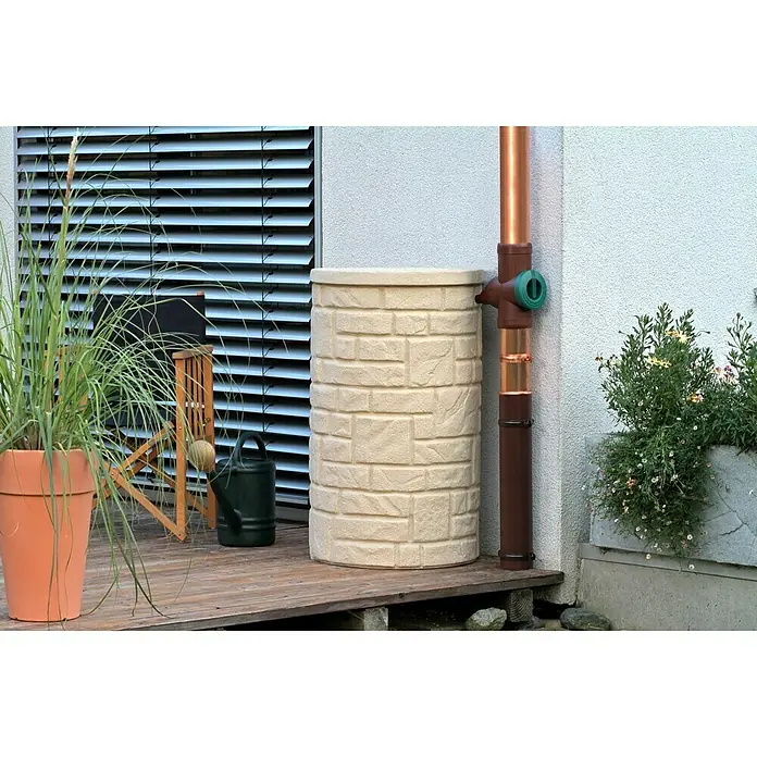 Arves Regenspeicher Arcado - 230 l, Sandsteinfarben, 1 Stk. - Aufnahme eines stilvollen Regenspeichers in Steinoptik auf einer Terrasse, neben einem Fallrohr und üppigem Grün.