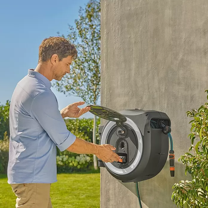 Gardena Power for All 18V Enrollador de manguera de batería Power-Roll XL - 18 V, Sin batería, Longitud del tubo: 35 m - Un hombre abre el portamangueras de pared gris oscuro y blanco, preparándose para usar o guardar la manguera de jardín.