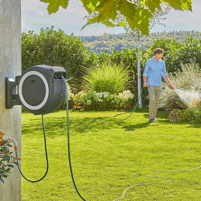 Gardena Power for All 18V Enrollador de manguera de batería Power-Roll XL - 18 V, Sin batería, Longitud del tubo: 35 m - Sistema automático de carrete de manguera gris y blanco, montado en pared, con manguera para un hombre que riega plantas en un jardín exuberante.