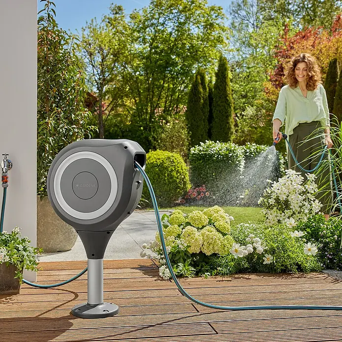 Gardena Schlauchbox Roll-Up S - Schlauchlänge: 15 m, 360 ° drehbar - Eine Gardena Schlauchtrommel mit Schlauch auf einer Holzterrasse, eine Frau bewässert im Hintergrund Pflanzen.