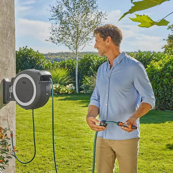 Gardena Power for All 18V Enrollador de manguera de batería Power-Roll XL - 18 V, Sin batería, Longitud del tubo: 35 m - Un hombre sostiene una manguera verde de un carrete de pared automático gris oscuro en un jardín.