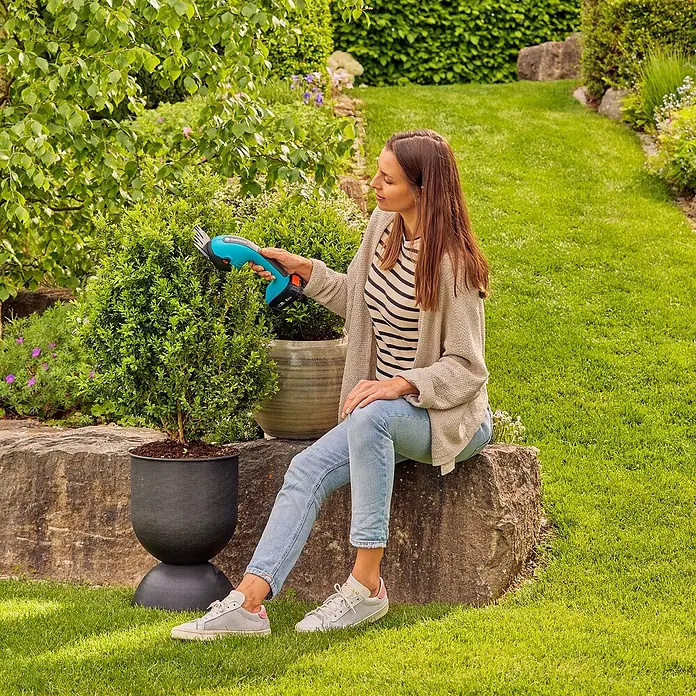 Gardena Power for All 18V Akku-Gras- & Strauchschere 20/18V P4A - 18 V, Li-Ionen, Ohne Akku - Eine Frau sitzt im Garten und schneidet mit einer Gardena Akku-Strauchschere präzise einen kleinen Busch. Der Garten ist gepflegt und idyllisch.