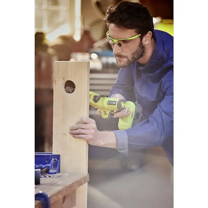 Ryobi ONE+ Akku-Bandschleifer R18PF-0 - 18 V, Ohne Akku - Ein Mann benutzt einen Ryobi Schleifer, um ein Holzstück zu bearbeiten, wobei er eine Schutzbrille trägt.