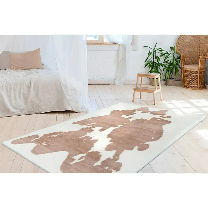 Kayoom Hochflorteppich Kuh - Weiß/Braun, 230 x 160 cm, 100 % Polyester - Ein weißer Teppich mit einem abstrakten, hellbraunen Muster liegt in einem hellen Raum mit einem Bett, einem Hocker und einem Rattansessel.