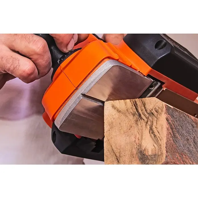 Black+Decker Cepilladora eléctrica BEW712 - 710 W, Profundidad de cepillo: 2 mm, Número de revoluciones de marcha en vacío: 16.500 r.p.m. - Primer plano de una cepilladora eléctrica de mano alisando una pieza de madera, mostrando los detalles naranjas y negros de la máquina para trabajar la madera.