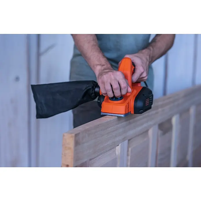 Black+Decker Cepilladora eléctrica BEW712 - 710 W, Profundidad de cepillo: 2 mm, Número de revoluciones de marcha en vacío: 16.500 r.p.m. - Primer plano de un hombre trabajando en un trozo de madera con una cepilladora, centrado en la cepilladora naranja.