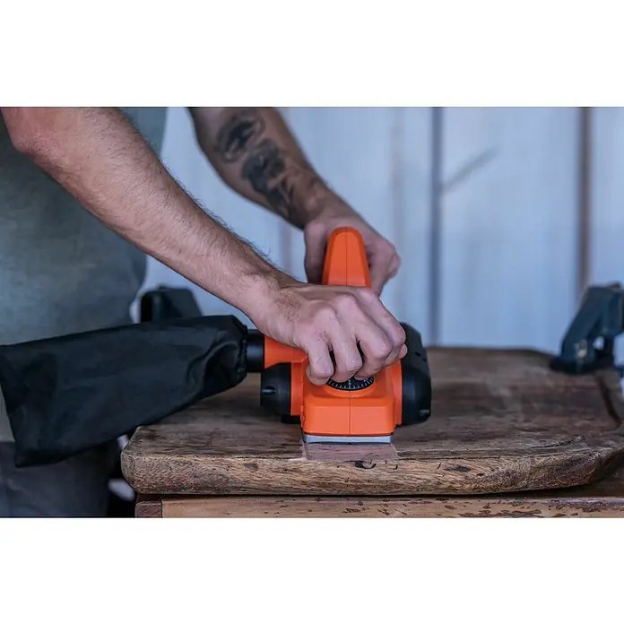 Black+Decker Cepilladora eléctrica BEW712 - 710 W, Profundidad de cepillo: 2 mm, Número de revoluciones de marcha en vacío: 16.500 r.p.m. - Imagen de un cepillo de carpintero naranja que está siendo utilizado por una persona sobre una mesa de madera para crear una superficie lisa.