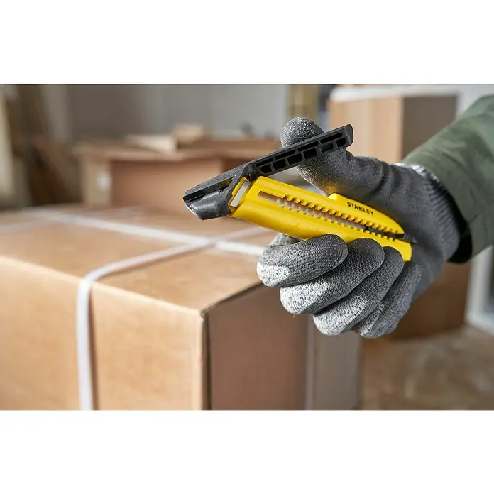 Stanley Cuchillo cúter STHT10489-0 - Plástico, Amarillo/Negro - Una mano con guantes de seguridad sostiene un cúter Stanley amarillo sobre una caja de cartón embalada, lista para abrirla.