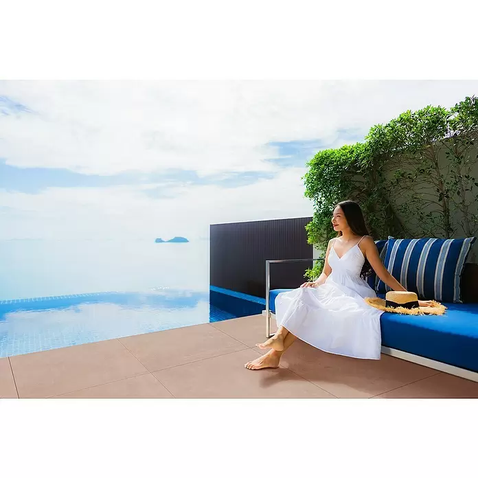 Terrassenfliese Modena - 80 x 80 x 2 cm, Taupe, Matt - Frau in weißem Kleid entspannt auf einem Outdoor-Sofa am Infinity-Pool mit Meerblick.
