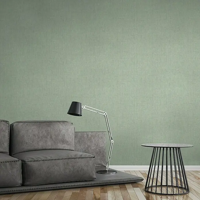 A.S. Création Greenery Vliestapete Borneo - Grün, Uni, 10,05 x 0,53 m - Ein Interieur mit einem grauen Sofa, einer Stehlampe und einem Beistelltisch vor einer grünen Wand. Die Lampe erhellt das Sofa auf einem Teppich.