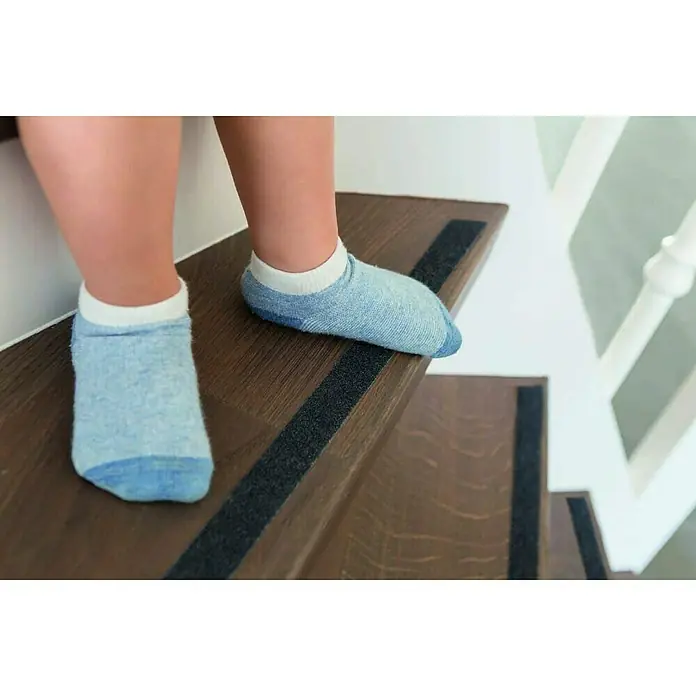 Abus Antirutschband Zoe - Schwarz, L x B: 400 x 2,4 cm - Aufnahme von einem Kind, das blaue Socken mit Antirutschsohle auf einer Holztreppe mit Antirutschstreifen trägt.
