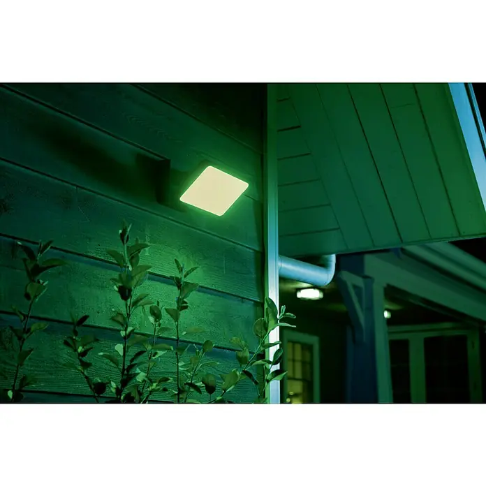 Philips Hue LED-Strahler White & Color Ambiance - 15 W, L x B x H: 22 x 16 x 15,3 cm, Schwarz, RGBW - Diese Nahaufnahme zeigt eine quadratische Philips Hue Außenleuchte mit grüner Beleuchtung, montiert an einer hölzernen Hauswand im Außenbereich.