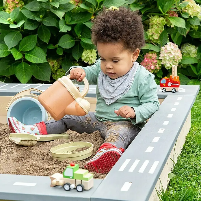 Muddy Buddy Sandkasten Highway Hero - L x B x H: 135 x 125 x 20 cm, Holz - Ein Kleinkind spielt in einem Sandkasten mit Straßendesign, umgeben von Sandspielzeug und einem Eimer.