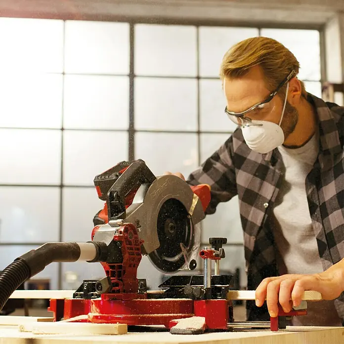Einhell Power X-Change 18V Akku-Kapp- & Gehrungssäge TE-MS 18-210 Li-Solo - 18 V, Ohne Akku, Durchmesser Sägeblatt: 210 mm, Leerlaufdrehzahl: 3.000 U/min - Ein Mann mit Schutzbrille und Staubmaske benutzt eine rote Einhell Kapp- und Gehrungssäge, um Holz zu schneiden. Staubpartikel sind in der Luft sichtbar.
