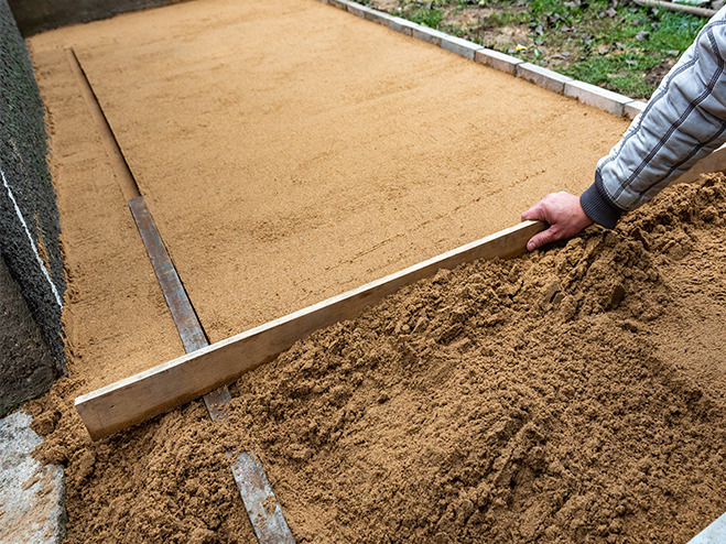 Mann verteilt mit einem Sand beim Pflastern als Unterbau mit Wasserwaage