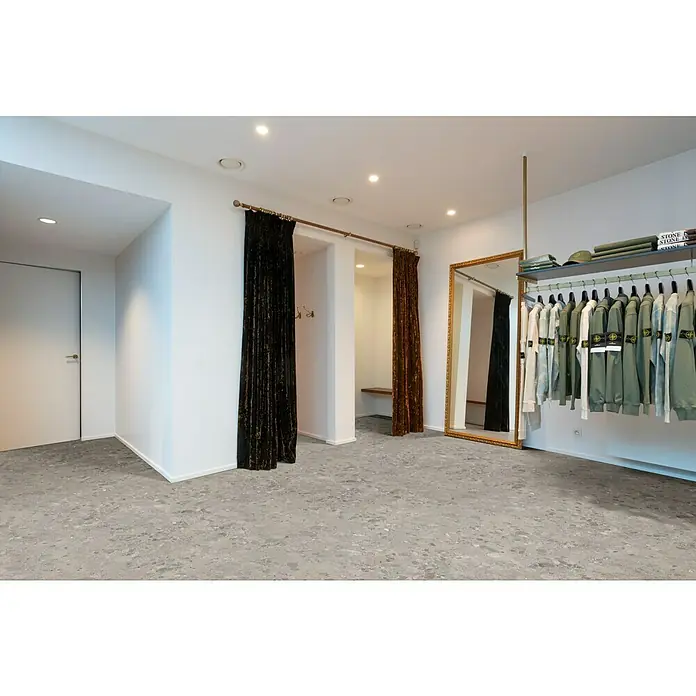 Beauflor PVC-Boden Meterware Hermes - Sonstige, Breite: 400 cm, Grau - Innenaufnahme eines Bekleidungsgeschäfts mit einer Kleiderstange voller Stone Island-Bekleidung, einem Spiegel und Umkleidekabinen.