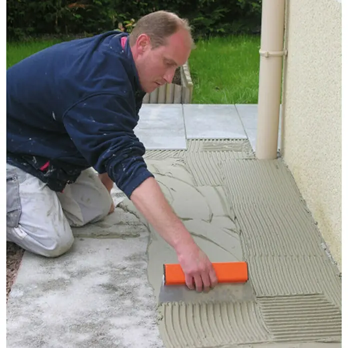 Mondelin Zahnspachtel - Zahnung: 10 mm, Klingenlänge: 300 mm - Ein Handwerker kniet und verteilt Fliesenkleber mit einer orangefarbenen Kelle auf einer Terrasse, um Fliesen zu verlegen.