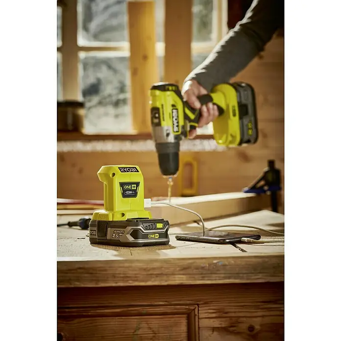 Ryobi ONE+ Adaptador USB R18USB - 18 V, Sin batería - Primer plano de un taladro inalámbrico RYOBI y un cargador en un banco de trabajo, con un teléfono inteligente cargándose a través del cargador.