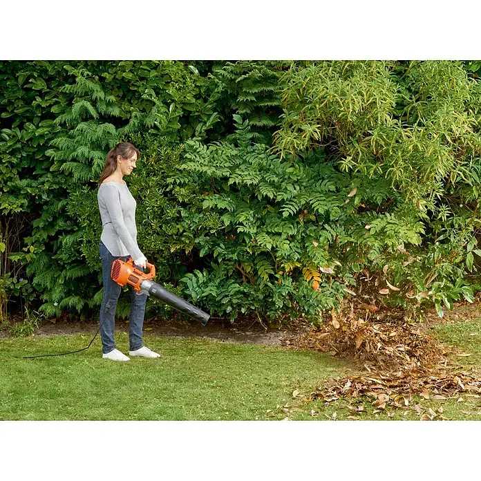 Black+Decker Soplador de hojas eléctrico BEBL 185-QS - 1.850 W, Velocidad de soplado: 225 km/h, Potencia de soplado: 12,7 m³/min - Mujer utilizando un soplador de hojas en el jardín para quitar las hojas. El soplador es naranja y negro.