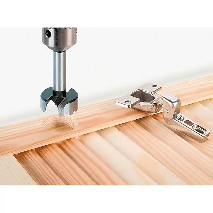 Wolfcraft Set de brocas Forstner - 5 pzs., Diámetro: 15 mm - 35 mm, Diámetro vástago: 10 mm - Broca de cazoleta metálica en madera, junto a una bisagra de mueble plateada, preparando un rebaje.