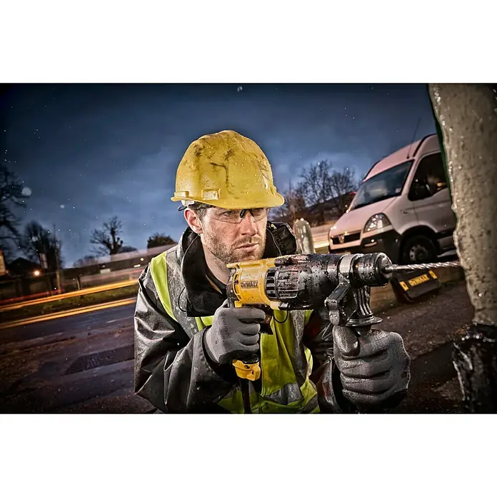 DeWalt Kombihammer D25133K - 800 W, 2,6 J, SDS-Plus-Aufnahme - Ein Bauarbeiter mit Schutzhelm und Dewalt-Bohrmaschine bohrt in eine Betonwand, während im Hintergrund ein Lieferwagen zu sehen ist.
