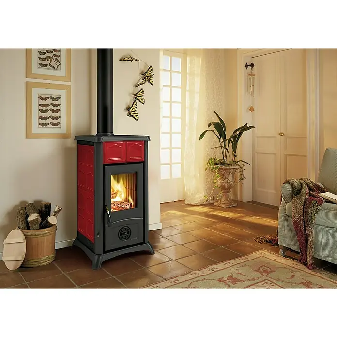 La Nordica Kaminofen Gemma - 7,3 kW, Raumheizvermögen: 172 m³, Material Verkleidung: Keramik, Schwarz/Bordeaux - Ein gemütliches Ambiente mit einem roten Kaminofen, der Wärme und Stil in den Raum bringt und eine behagliche Atmosphäre schafft.