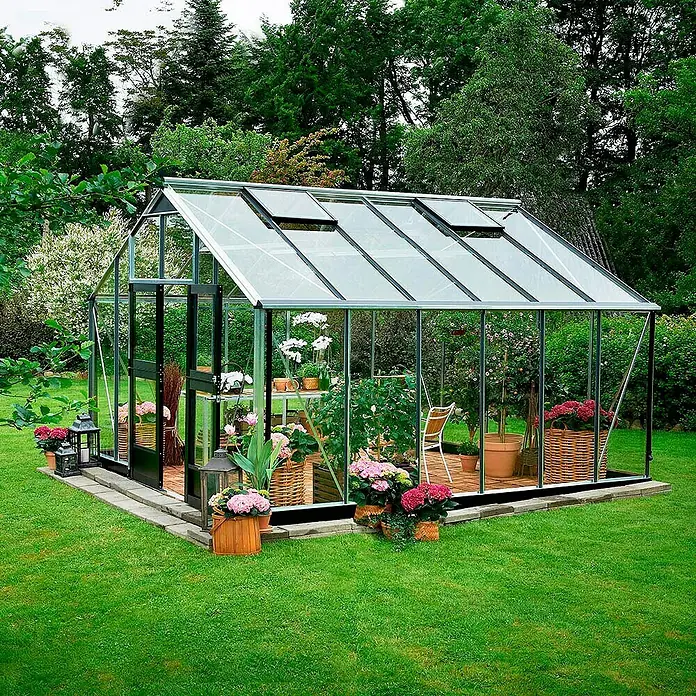 Juliana Gewächshaus Gärtner - Außenmaß inkl. Dachüberstand (B x T): 368 x 439 cm, Material Seitenwand: Glas, 3 mm, Silber/Schwarz - Das Bild zeigt ein Gewächshaus mit Blumen und Pflanzen, das auf einem Rasen steht und von Bäumen umgeben ist.