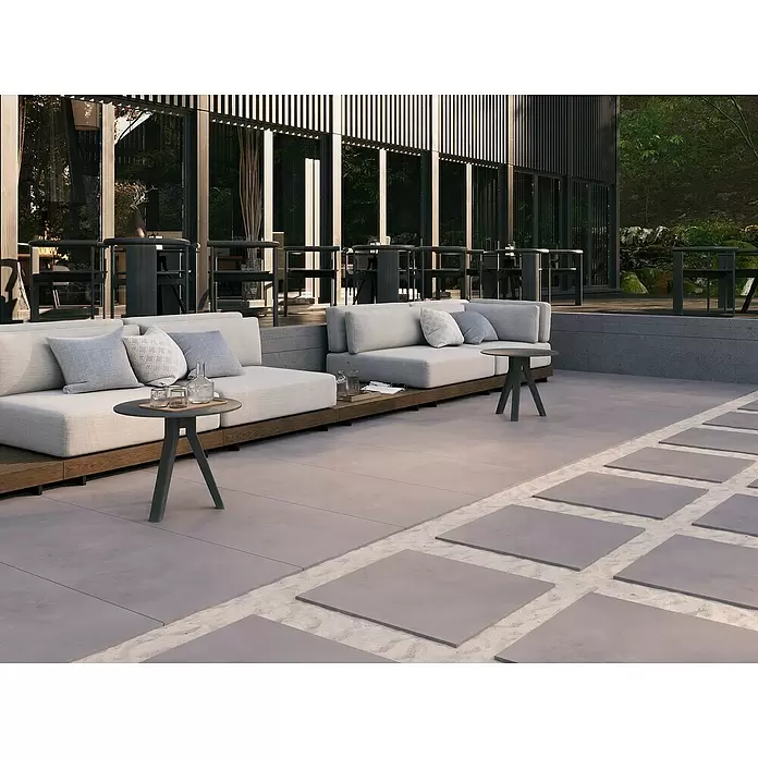 Terrassenfliese Modena - 60 x 60 x 2 cm, Ash/Graubraun, Matt - Moderne Outdoor-Lounge-Sitzgruppe mit hellgrauen Kissen und Holzsockeln, kombiniert mit dunklen Beistelltischen auf einer modernen Terrasse.