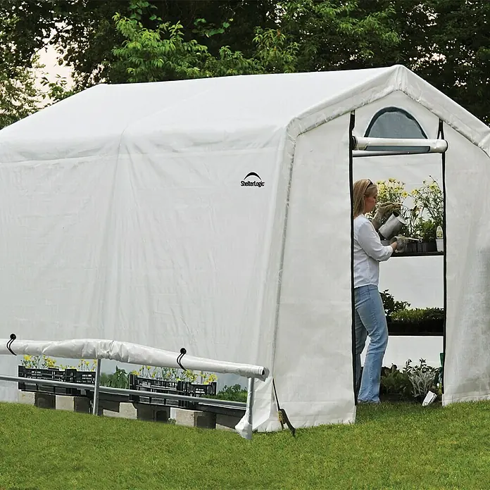 ShelterLogic Gewächshaus - Außenmaß inkl. Dachüberstand (B x T): 240 x 240 cm, Grau, Grundfläche: 5,76 m² - Ein ShelterLogic-Gewächshaus mit einer Frau, die Pflanzen gießt, und einer großen Auswahl an Pflanzen, die zum Verkauf bereitstehen.
