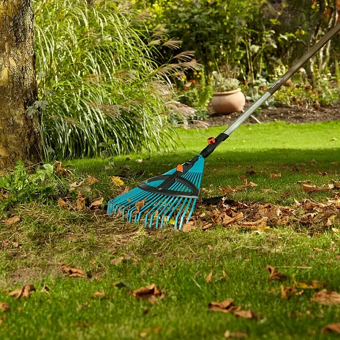 Gardena Combisystem Laubbesen - 35 cm - 52 cm, Mit Stiel - Nahaufnahme eines blauen Laubrechens der Marke Gardena, der auf einem Rasen mit Herbstlaub liegt, im Hintergrund ein Baumstamm und Pflanzen.