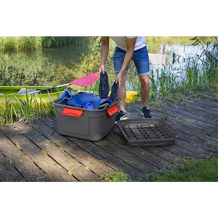 Sunware Aufbewahrungsbox Q-Line wasserdicht - L x B x H: 60,1 x 53,1 x 42,5 cm, Kunststoff, Anthrazit - Ein Mann packt Neoprenschuhe in eine graue Aufbewahrungsbox mit rotem Verschluss neben einem gelben Kajak am Ufer.
