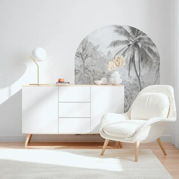 Vinilo de pared autoadhesivo Arco Tropic Dune - 132 x 170 cm, Blanco/Negro, Plástico - Un interior elegante con un aparador blanco, una lámpara y un sillón cómodo sobre una alfombra. Un mural decorativo completa el aspecto.