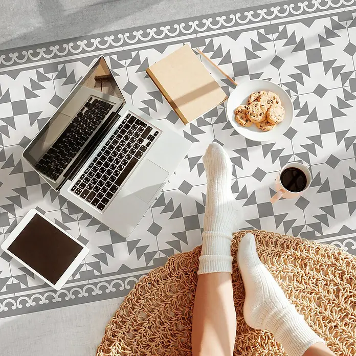 Alfombra de vinilo Buzz - Gris, 200 x 60 cm, PVC - Portátil, tableta, cuaderno, galletas y café sobre superficie estampada, con piernas en calcetines blancos descansando sobre una alfombra redonda.