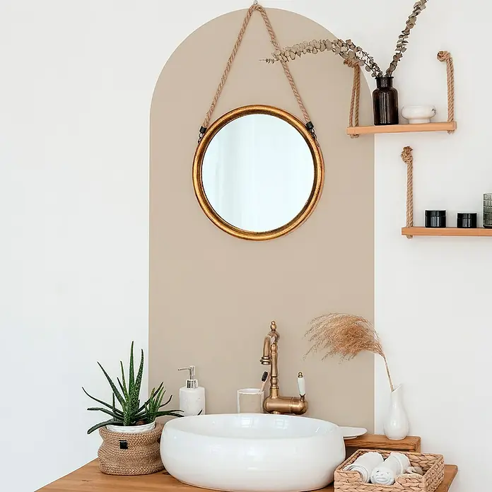 Vinilo de pared autoadhesivo Arco Topo - 65 x 200 cm, Topo, Plástico - Un tocador de baño con un espejo redondo con marco dorado que cuelga de una cuerda, con estantes decorativos, dispensador de jabón y vegetación.
