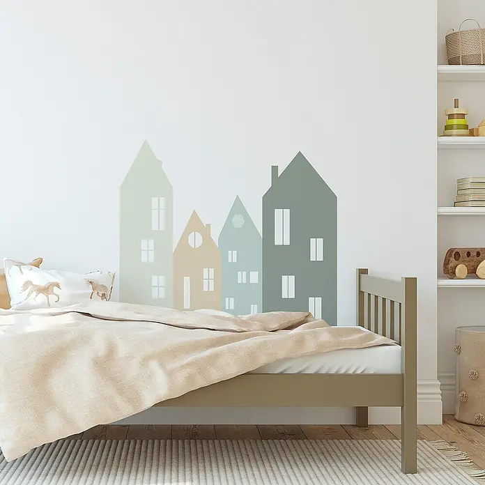 Vinilo de pared autoadhesivo Arco Green Houses - 132 x 170 cm, Multicolor, Plástico - Habitación infantil con una cama baja y una decoración de pared de casas en tonos pastel. Colores claros y ambiente acogedor.