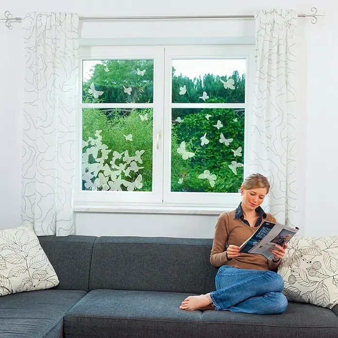 Komar Fenstersticker Schmetterlinge - Weiß, 31 x 31 cm, 21 -tlg. - Eine Frau sitzt auf einem Sofa und liest eine Zeitschrift vor einem Fenster mit Schmetterlingsaufklebern.