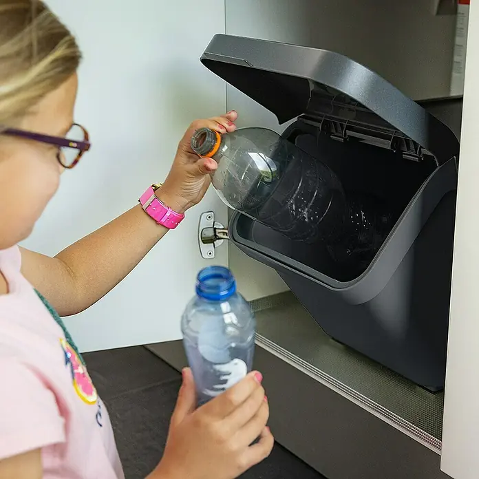 Sunware Mülltrennsystem Sigma Home - L x B x H: 49,3 x 27 x 29,8 cm, Kunststoff, Anthrazit - Ein Kind wirft eine Plastikflasche in einen grauen Recycling-Behälter. Der Behälter ist in einem Schrank verbaut.